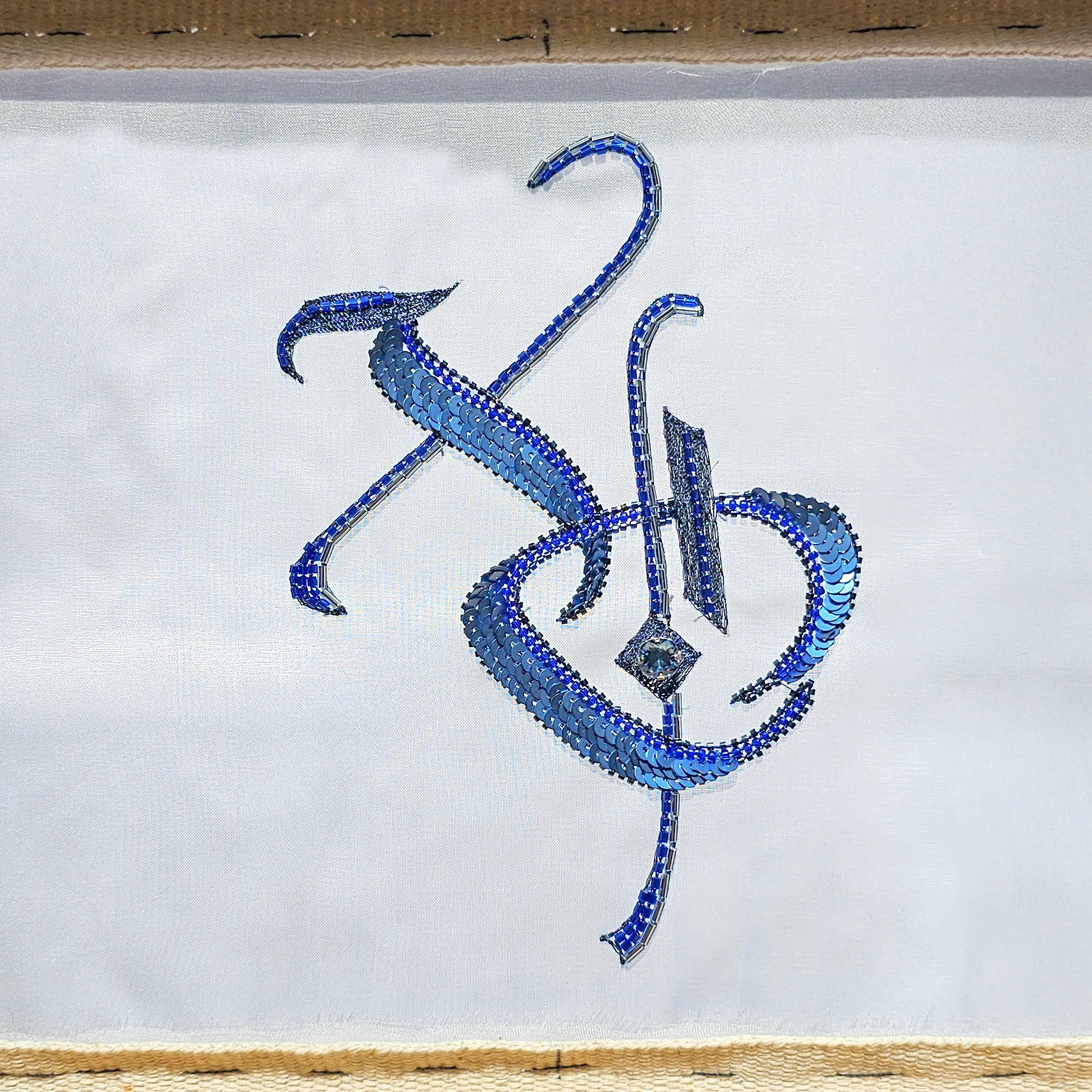 XO monogram embroidered with blue sequins and beads using Luneville embroidery, a couture technique taught step by step for beginners, stitched on white sheer fabric and shown on an embroidery frame