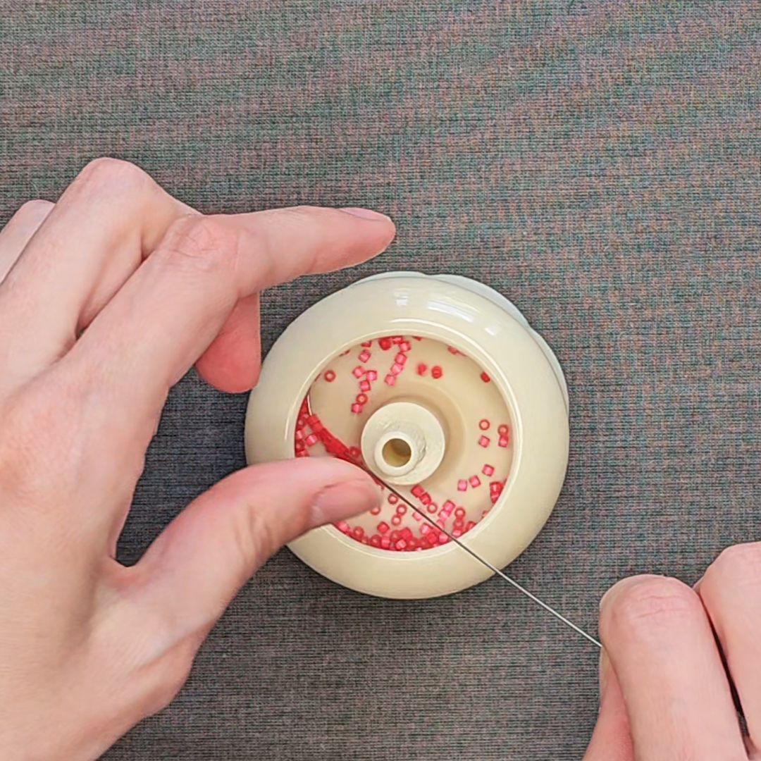 Close-up of hands using a curved needle with a plastic manual bead spinner to string red seed beads for embroidery preparation.