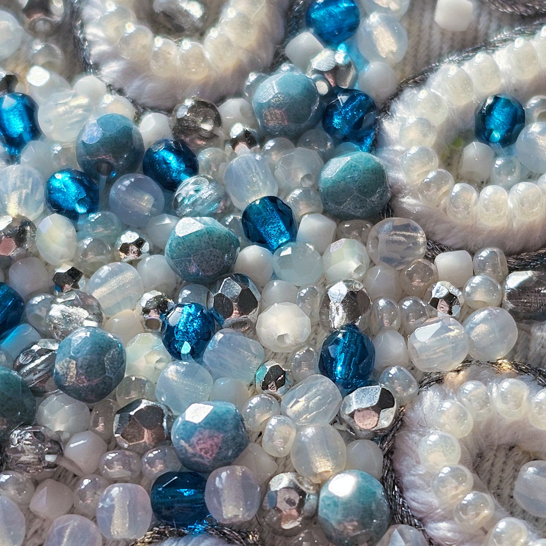 Macro photograph showing the detailed surface of the Jewels of Winter embroidery design. Blue, white, translucent and silver beads are combined with pearl embroidery and metallic elements to create variation in colour, texture and reflection.