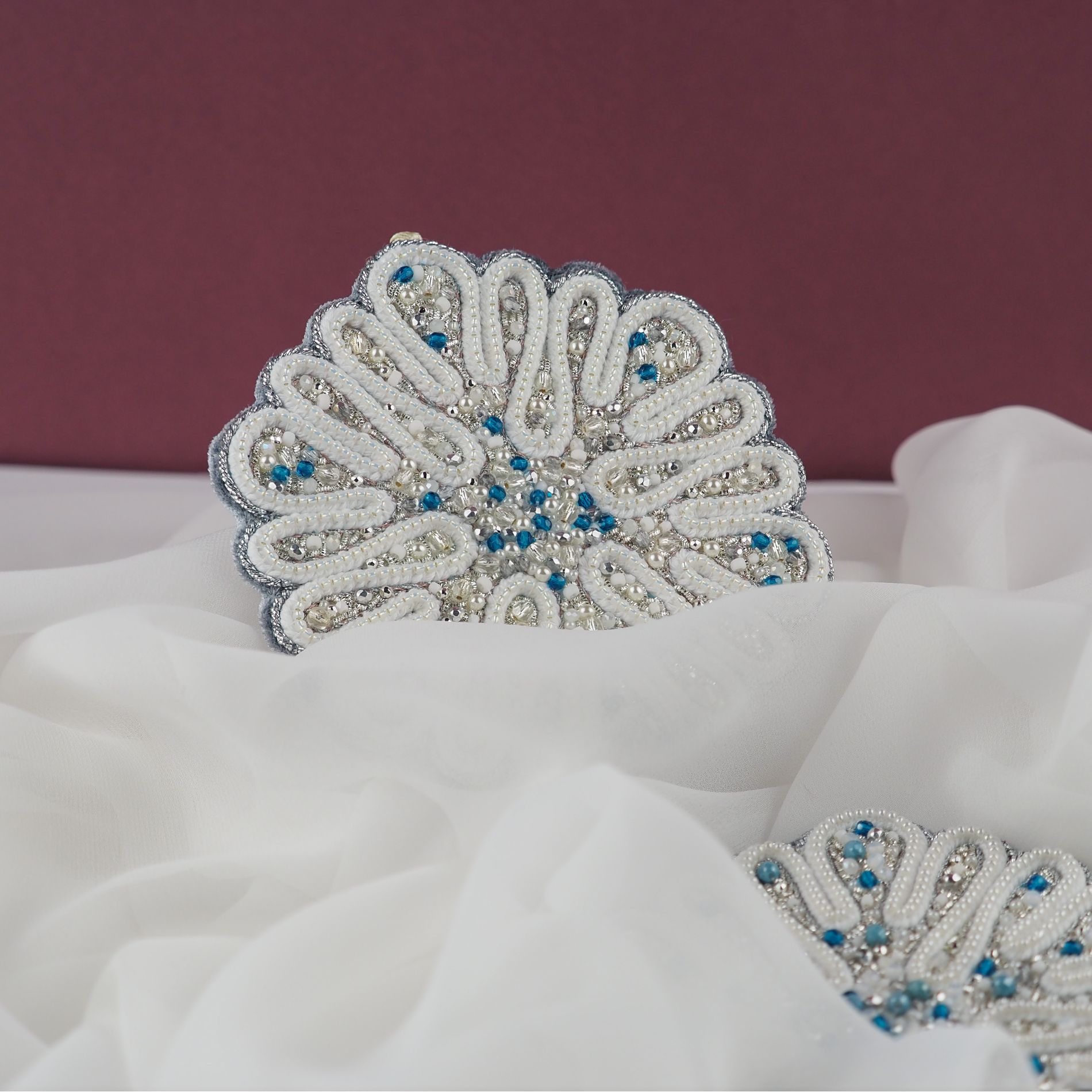 Two completed textile ornaments from the Jewels of Winter course, photographed among soft white fabric. Each piece is created by hand using pearl embroidery, beads, crystals and metallic details in a winter-inspired palette.