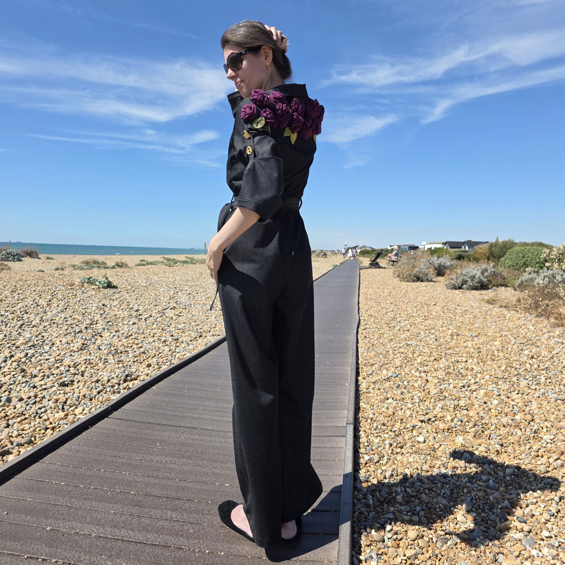 Three-quarter view of a black jumpsuit with purple fabric roses and embroidered leaves extending across one shoulder and the back.