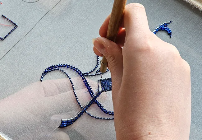 Hand using a Luneville hook to embroider a curved design with blue seed beads and bugle beads on sheer fabric.
