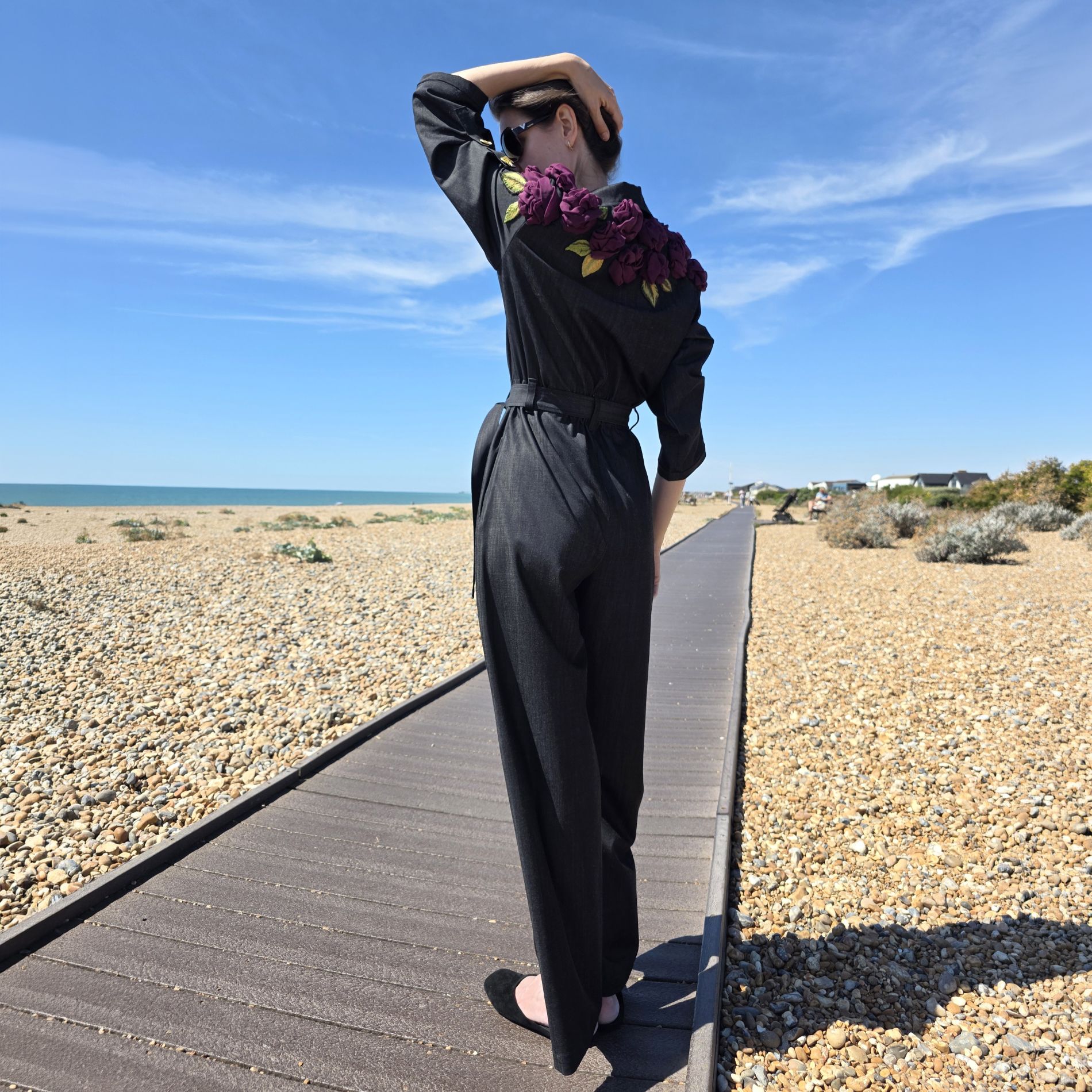 Full-length back view of the black Rose Jumpsuit with handmade deep purple fabric roses and embroidered leaves positioned across the upper back and shoulders. The image shows the scale of the floral composition in relation to the complete garment.