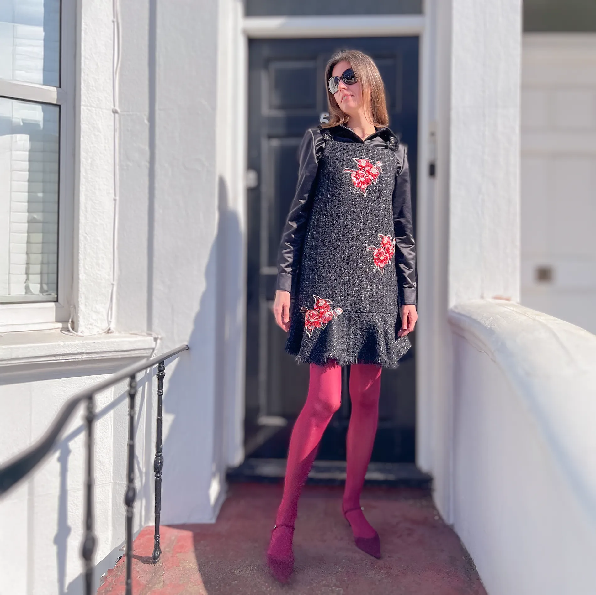 A woman poses on a white townhouse porch wearing a black tweed dress adorned with red camellia embroidery, created using techniques from the Couture Camellias course. Styled with burgundy tights, matching heels, and a black long-sleeve top underneath, the outfit demonstrates how to apply course skills to fashion pieces.